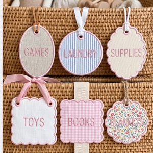 May include: Set of six decorative storage tags with embroidered text. The tags have various shapes, including oval, round, and scalloped edges. The tags feature the words "GAMES", "LAUNDRY", "SUPPLIES", "TOYS", "BOOKS", and "BLANKETS" in pink thread.