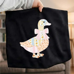 May include: A black t-shirt featuring a central graphic of a duck composed of various pastel-coloured floral and plaid fabric patterns. The duck is depicted in profile, facing right, and wears a large, decorative bow around its neck in a matching style.
