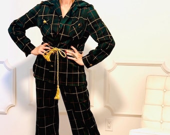 May include: A dark green plaid suit with gold accents. The suit includes a blazer and wide-leg pants. A gold rope belt with a tassel cinches the waist. Gold sandals complete the outfit. The background is a white wall with decorative molding.