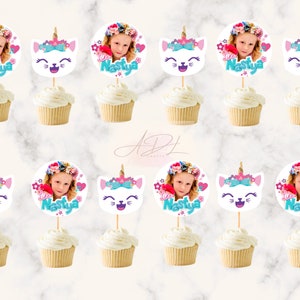 May include: Twelve cupcake toppers featuring a white cat face with a unicorn horn, pink flowers, and a blue bow. Each topper also includes a circle with a girl's photo and the name "Nastya" in pink and blue lettering.