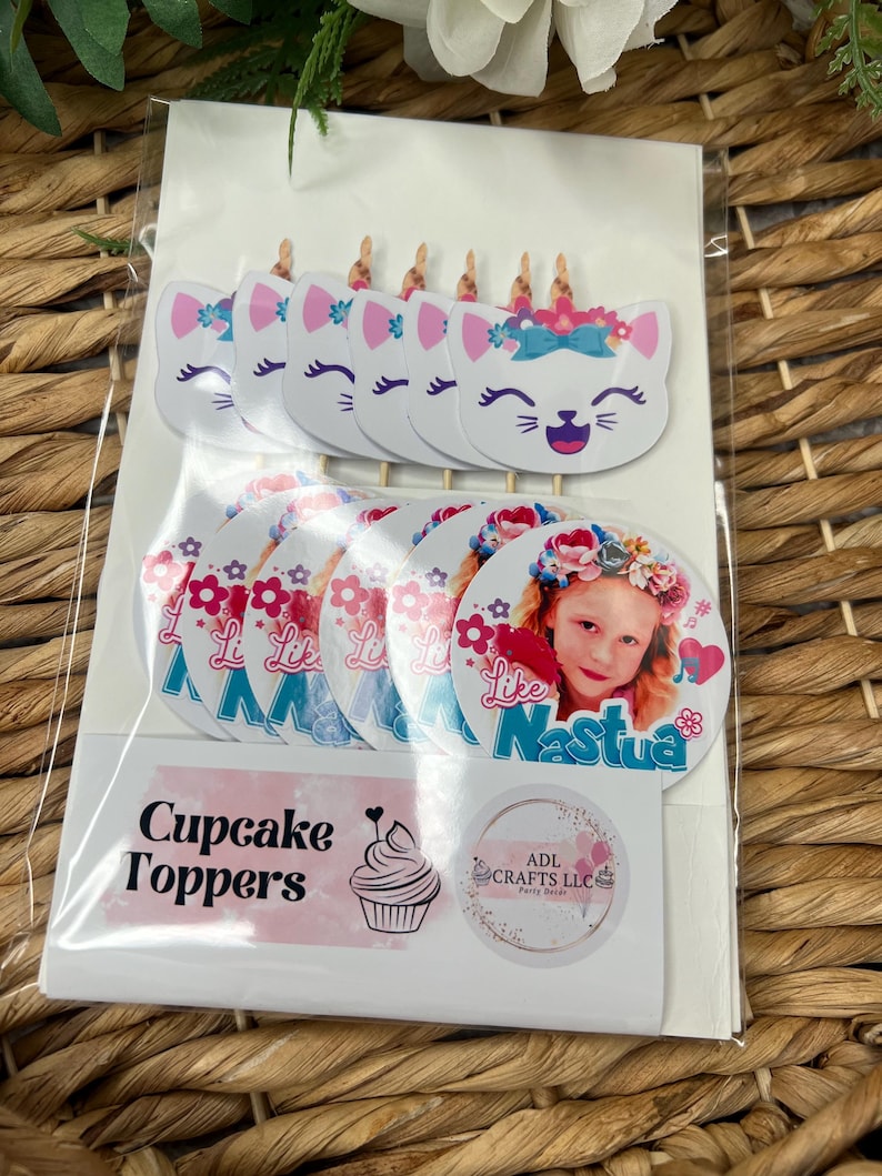 May include: A set of cupcake toppers featuring a white cat with a pink bow and blue flowers, and a round topper with a girl's photo and the text "Like Nastya". The set also includes a sticker with the text "Cupcake Toppers" and a cupcake graphic, and a sticker with the text "ADL Crafts LLC".