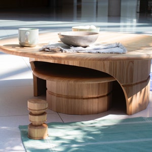 May include: A wooden coffee table with a unique design featuring a curved lower shelf and a round top. The table is made of light wood and has a natural finish. There are two bowls and a mug on the table, and a small wooden stool is on the floor next to the table.