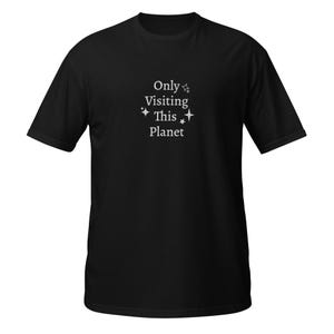 Pode incluir: T-shirt preto com o texto branco "Only Visiting This Planet" e estrelas. A camiseta tem gola redonda e parece ser feita de um material macio.