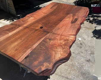 May include: A large, rustic wooden table top with a live edge. The wood is a rich brown color with a natural grain pattern. The table top is smooth and polished.