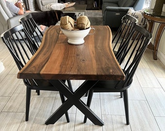 Puede incluir: Una mesa de comedor de madera de borde vivo con una base en forma de X negra y cuatro sillas negras. La mesa tiene un acabado de madera natural y está rodeada de un suelo de baldosas de color gris claro. Un perro está sentado en primer plano.