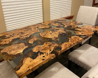 May include: A large, rustic wooden dining table with a clear epoxy resin top. The table has a natural wood grain pattern and features a unique, swirling design in the resin. The table is surrounded by four chairs with light gray fabric upholstery.