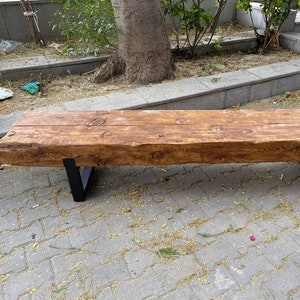 May include: A rustic wooden bench with a natural wood finish and black metal legs. The bench is made from a single piece of wood and has a rough, textured surface.