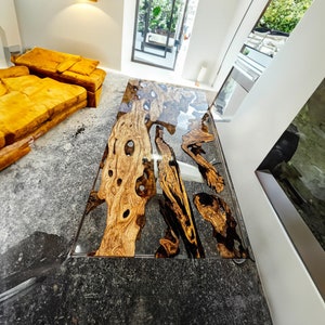 May include: A large, rectangular table made of wood and epoxy resin. The wood has a natural, rustic look with knots and grain patterns. The epoxy resin is clear and creates a smooth, glossy finish. The table is sitting on a gray concrete floor.