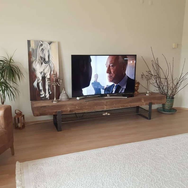 May include: A rustic wooden TV stand with a metal frame and a black metal shelf. The stand is in a living room setting with a flat screen TV, a painting of a white horse, and a potted plant.