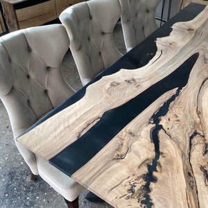 May include: A large wooden dining table with a black epoxy resin inlay. The table has a natural wood grain pattern and is surrounded by four chairs with tufted upholstery.