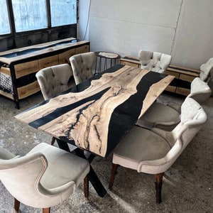 May include: A large wooden dining table with a black epoxy resin river design. The table has a black metal X-shaped base and is surrounded by six beige upholstered chairs.