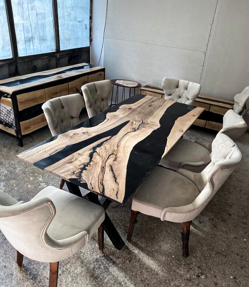 May include: A large wooden dining table with a black epoxy resin river design. The table has a black metal X-shaped base and is surrounded by six beige upholstered chairs.
