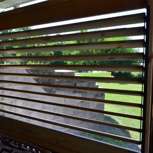 May include: Wooden slatted window with a view of a grassy area and stone structure. The window is made of horizontal wooden slats, allowing light and air to pass through. The frame is made of wood.