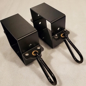 May include: Two matte black metal brackets with rectangular frames, each featuring a circular mounting hole on the side and a small metal plate attached with two silver screws. A gold-toned grommet holds a black carabiner clip on each bracket.
