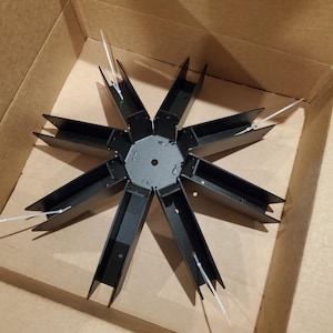 May include: A black metal star-shaped component with eight arms, each featuring a rectangular channel. White cable ties secure the arms. The object is positioned inside a cardboard box, possibly a part of a larger assembly.