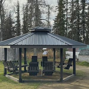 Puede incluir: Un cenador gris oscuro con techo de metal y estructura de madera. El cenador tiene varias sillas y columpios de madera en su interior. La estructura está situada en una zona de césped con árboles al fondo.