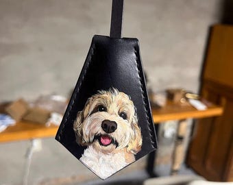 May include: Black leather key fob with a hand-painted portrait of a fluffy, light brown and white dog. The dog has a happy expression with its tongue sticking out. The fob has a black strap attached to the top.