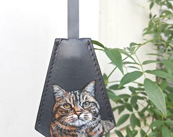 May include: A black leather key fob with a hand-painted portrait of a tabby cat. The cat has brown and gray stripes, with one yellow and one blue eye. A gray strap is attached to the top of the fob.