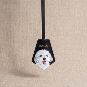 May include: A black leather luggage tag with a pointed bottom edge and a vertical strap. The tag features a hand-painted portrait of a white fluffy dog with a pink tongue sticking out and the name GORDO printed in gold letters above the dog.