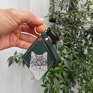 A hand is holding a green keychain with a picture of a cat on it, against a backdrop of greenery.