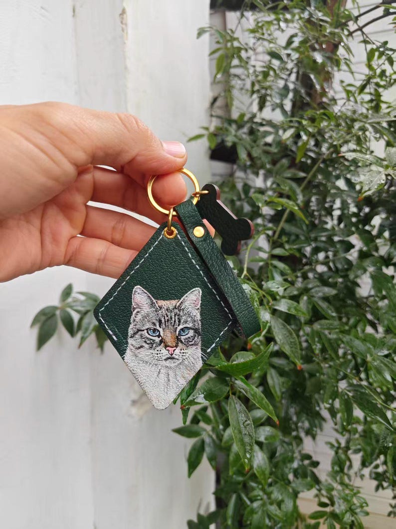 A hand is holding a green keychain with a picture of a cat on it, against a backdrop of greenery.