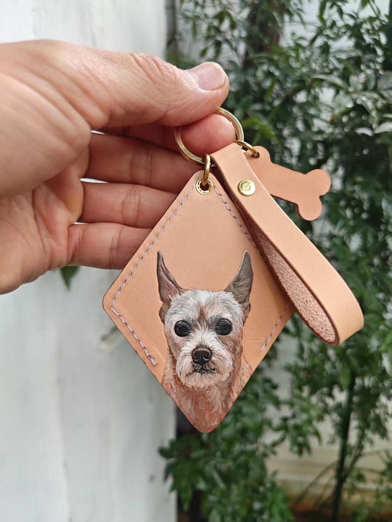 A hand is holding a keychain with a picture of a dog on it, against a backdrop of greenery.