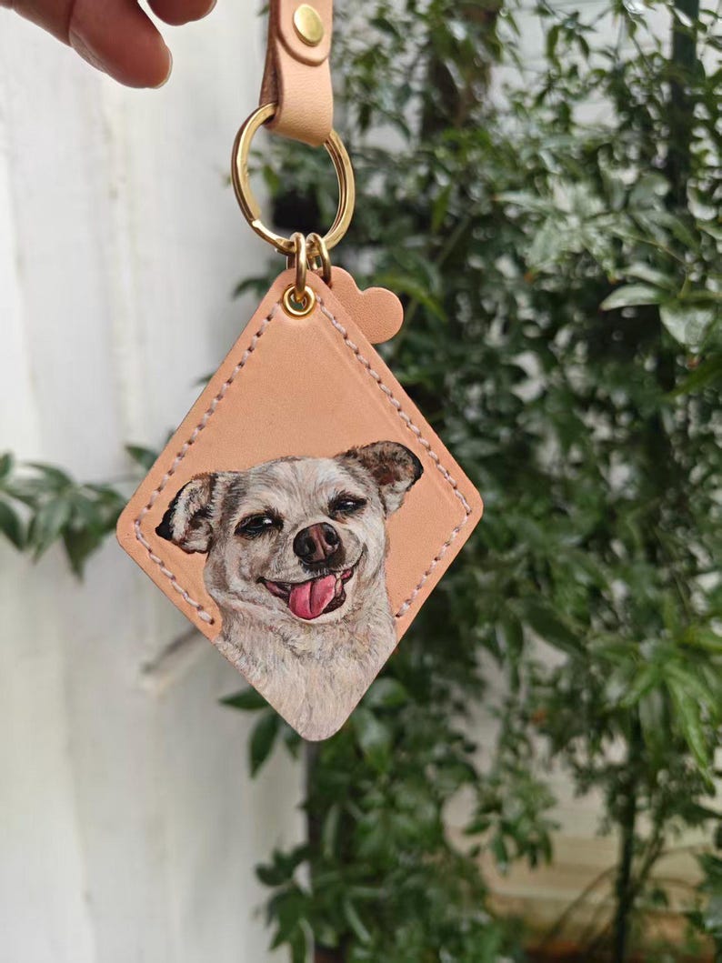 A hand is holding a keychain with a picture of a dog on it, set against a backdrop of greenery.