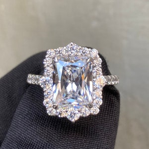 Puede incluir: Un anillo plateado con una gran piedra preciosa rectangular y transparente en el centro, rodeada de piedras redondas y transparentes más pequeñas. El anillo está sobre una superficie negra, con un fondo neutro.