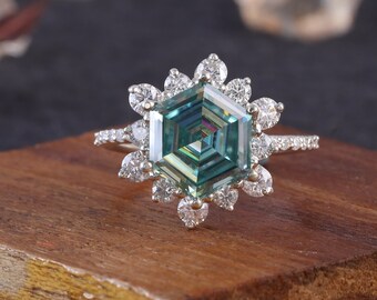 May include: A hexagonal green moissanite engagement ring with a halo of smaller diamonds set in a white gold band. The ring is displayed on a wooden surface.