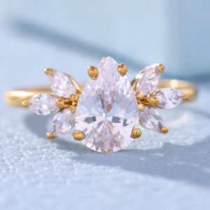 Könnte beinhalten: Ein Verlobungsring mit einem birnenförmigen Diamanten, einem goldenen Band und kleineren Diamanten, die in einem floralen Design gesetzt sind.