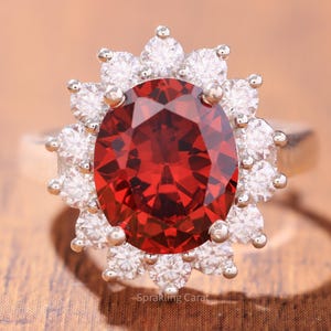 May include: A silver ring featuring a large, oval-cut, deep red gemstone surrounded by a halo of smaller, round, white gemstones. The ring is set against a wooden surface, with the text "Sprakling Carat" visible.