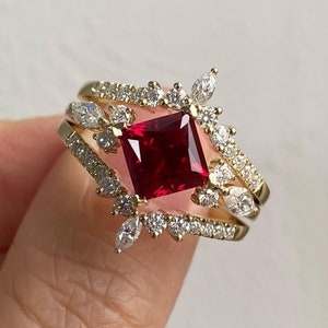 Könnte beinhalten: Ein goldener Ring mit einem großen, quadratischen Rubin-Edelstein, der von kleineren Diamanten umgeben ist. Der Ring hat ein zartes, florales Design mit kleinen Diamant-Akzenten.