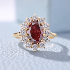 May include: A gold ring featuring a large, oval-cut, deep red gemstone surrounded by a halo of smaller, round, white gemstones. The ring's band is a simple, polished gold. The setting of the central stone is also gold.
