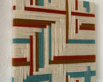 May include: A wall hanging made of woven yarn in shades of beige, brown, orange, and teal. The yarn is woven in a geometric pattern, creating a modern and abstract design.