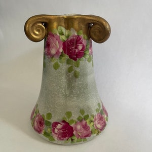 Op de afbeelding: Een kleine, witte keramische vaas met een groen en roze bloemenpatroon. De vaas heeft twee gouden, gerolde handvatten.