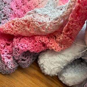 May include: A crocheted blanket featuring a gradient color pattern that transitions from light gray to soft pink and vibrant magenta. The textile has a scalloped edge design with a textured, open-work stitch pattern and is paired with a plush white fabric.