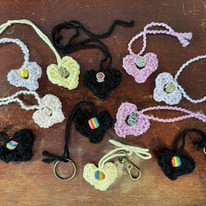 Peut inclure: Collection de pendentifs et porte-clés en forme de cœur crochetés à la main, de différentes couleurs, dont le noir, le lavande et le jaune. Certains cœurs présentent des ornements décoratifs et des accents arc-en-ciel. Les porte-clés ont des fermoirs en métal.