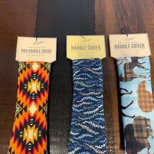 May include: Three handmade pan handle covers. The first is orange, black, and white with a geometric pattern. The second is blue with a white floral pattern. The third is blue with a brown and black plaid pattern.