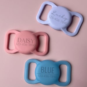 May include: Three personalised dog collar buckle add-ons. The first is light blue with the name "Lucky" and phone number "123-456-7890". The second is pink with the name "Daisy" and phone number "123-456-7890". The third is light blue with the name "Blue" and phone number "123-456-7890".
