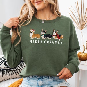 Może przedstawiać: Zielony sweter z świątecznym wzorem przedstawiającym trzy corgi w czapkach Mikołaja i szalikach. Pod corgiami znajduje się napis "Merry Corgmas".
