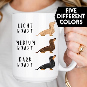 May include: White ceramic coffee mug featuring a dachshund illustration for each roast: light, medium, and dark. The text "Five Different Colours" is on a black rectangle.