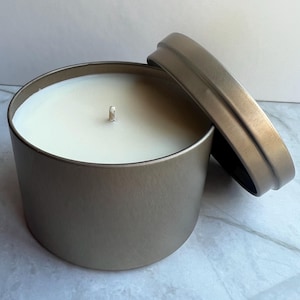May include: A round, metallic candle tin with a lid slightly ajar. The candle inside is white with a central wick. The tin has a muted gold color, and the lid rests beside the open container. The candle is a home decor item.