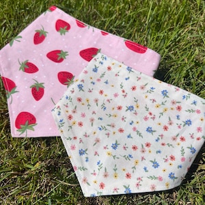 May include: Two bandanas with different patterns are displayed on green grass. One is pink with red strawberries and white polka dots. The other is white with a floral pattern of small blue, yellow, and pink flowers.