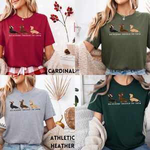 May include: A t-shirt with a dachshund wearing a Santa hat and the text "Dachshund Through The Snow". The shirt is available in five colors: cardinal, military green, athletic heather, forest green, and black.