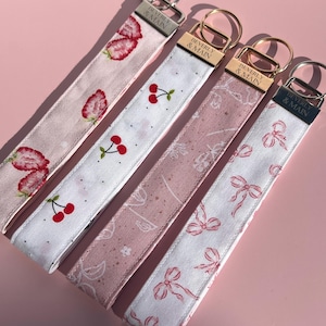 May include: Four fabric keychains with different patterns: strawberries, cherries, floral, and bows. Each keychain has a silver or gold-colored metal clasp and a tag that reads "BEVERLY & MAIN". The keychains are in shades of pink and white.