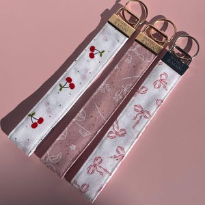 Puede incluir: Cuatro llaveros de tela con herrajes dorados y el texto "BEVERLY & MAIN". Los llaveros presentan varios diseños: cerezas sobre blanco, flores sobre rosa y lazos sobre blanco. Los llaveros están sobre una superficie rosa.