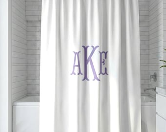 May include: A white shower curtain featuring a large, centered monogram with the letters AKE in a light purple, serif font. The curtain hangs from a silver metal rod and displays vertical folds throughout the fabric, creating a clean look.
