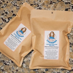 May include: Two brown paper bags with a label that says "TEENWAH QUINOA PROTEIN BAR MIX Made In A Home Kitchen" and a cartoon illustration of a woman. The label also includes the ingredients and a warning that the product contains peanuts, walnuts, and coconut.