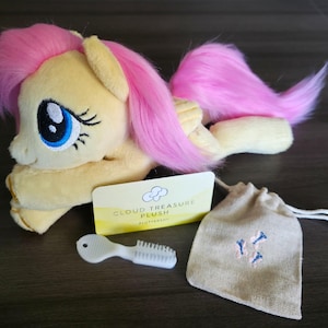 May include: A yellow plush pony with pink hair and a matching tail. The image includes a brush, a drawstring bag with embroidered details, and a card that reads "CLOUD TREASURE PLUSH".