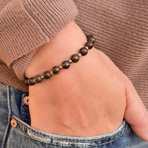 Op de afbeelding: Een armband met donkere, ronde kralen en goudkleurige tussenstukken. De armband wordt om een pols gedragen, met een bruine trui en een blauwe spijkerbroek op de achtergrond. De kralen zijn ongeveer 0,8 cm in diameter.